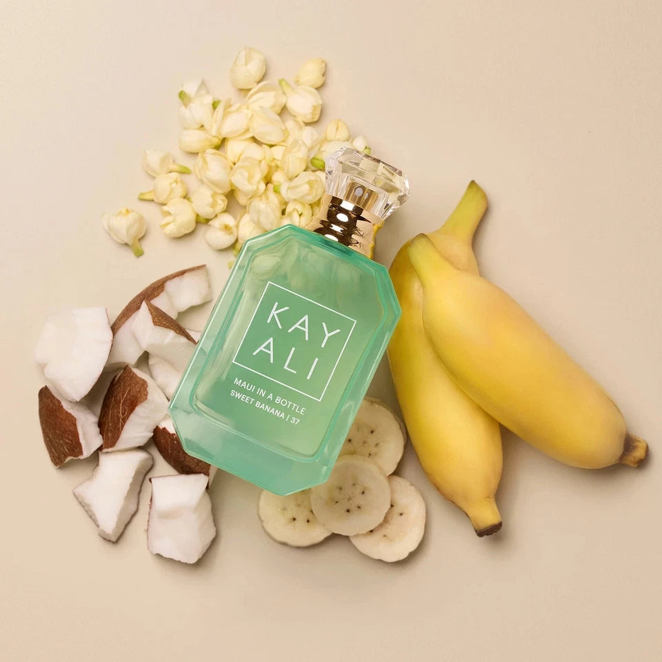 Perfume bottle labeled 'KAY ALI' surrounded by bananas, coconuts, and flowers on a beige background